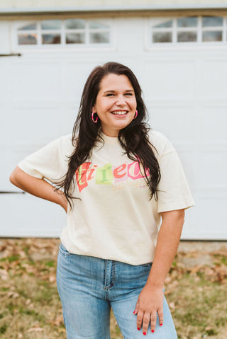 A woman smiling outdoors in front of a white garage door, wearing a cream graphic t-shirt with a colorful 'Fried' text design and light blue jeans.