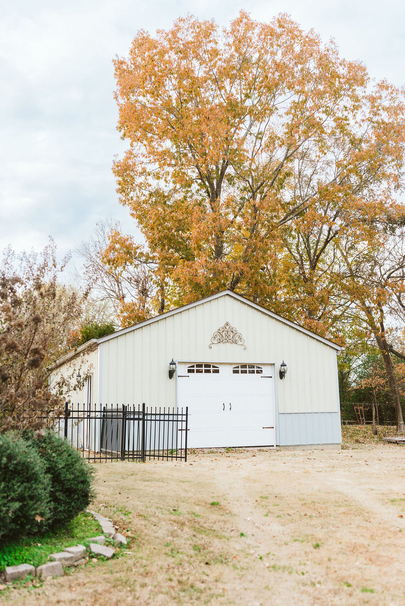 A white metal barn-style building with a large white garage door, set in a yard with dry grass, shrubs, and tall trees in autumn colors.