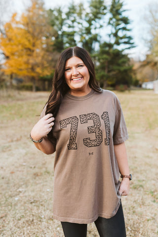 brunette female model wearing a mocha brown color short-sleeve graphic t-shirt with a printed design in black ink that reads "731" with a leopard animal print insdie the font