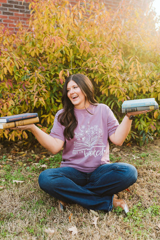 female model modeling a purple comfort colors short-sleeve tee with a graphic print that reads "sorry, I'm booked" with a floral print with an open book. she's also holding books in her hands as props.