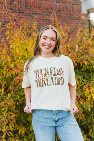 female model wearing an ivory graphic tee that reads "tucked & threaded" in a camouflage print