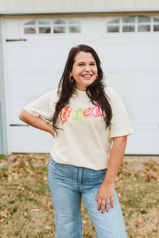 ivory color short-sleeve graphic t-shirt with a printed design in green, red, yellow, and purple ink that reads "tired, but trying" in a collage style print